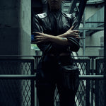 Lily Gatins wearing the black SYZMIC JACKET EIDOS-408, standing with crossed arms in an industrial setting. Photography by Nox / Lux.