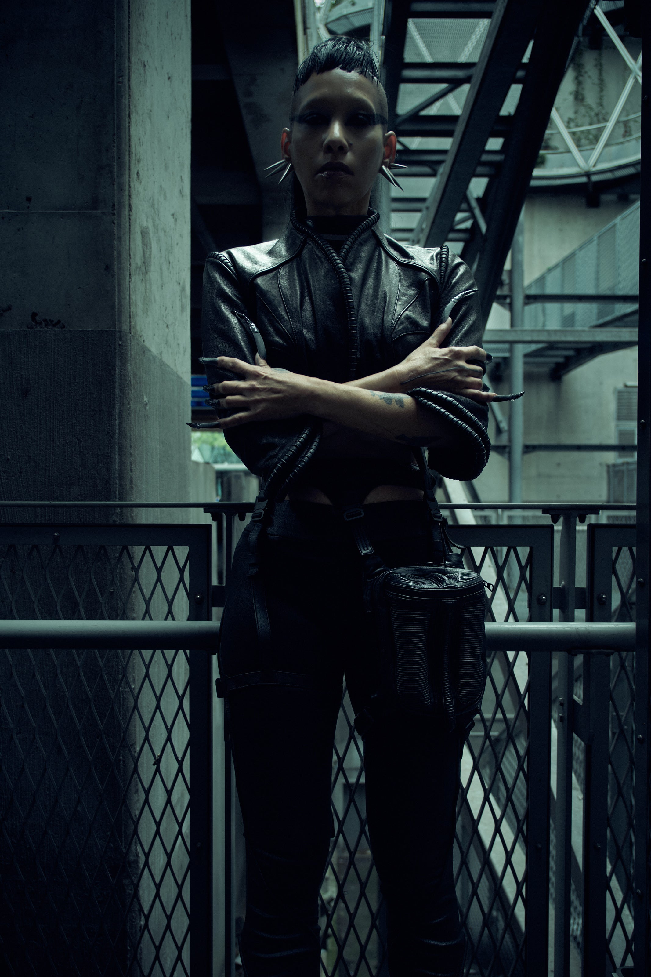 Lily Gatins wearing the black SYZMIC JACKET EIDOS-408, standing with crossed arms in an industrial setting. Photography by Nox / Lux.